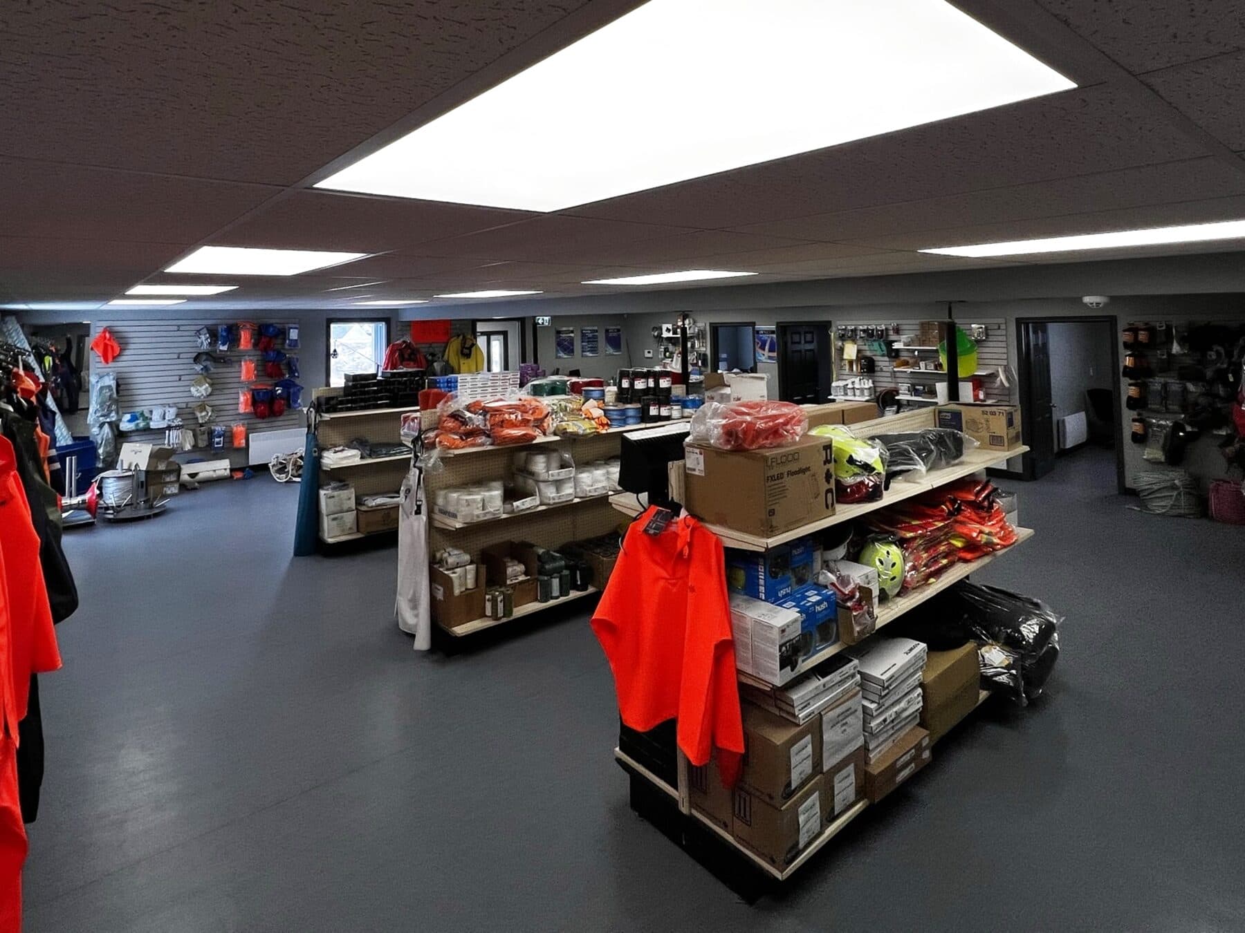 The Port au Choix store floor — orange rain gear and flotation jackets on the rail, hard hats and packaged gear on the shelving, and a wall of shackles, hooks and master links to the right