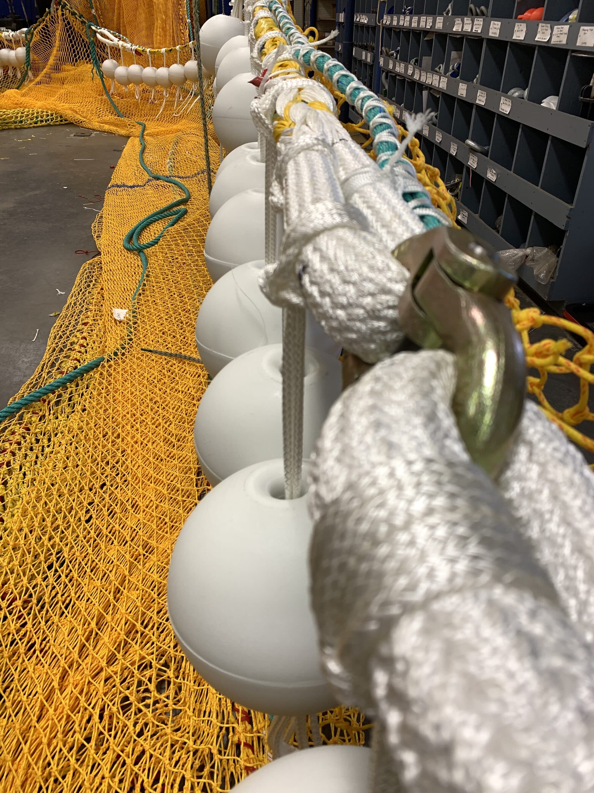 A float line on the shop floor, white floats threaded on white braided rope with a galvanised shackle joining the run, yellow netting laid out behind