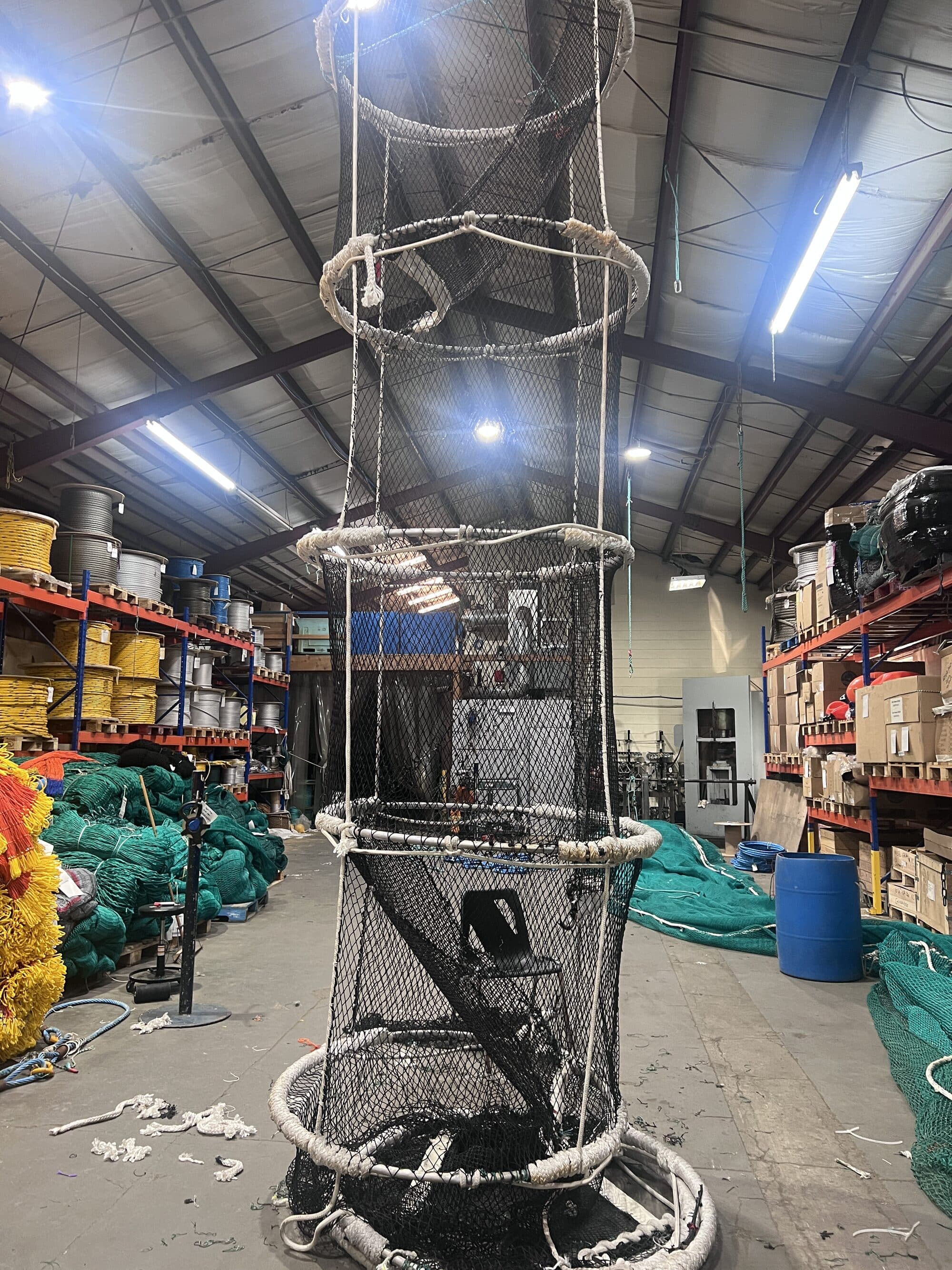 A tall cylindrical netted enclosure hung from a hoist on the shop floor, dark mesh laced to three white-roped hoops, netting stock and rope reels behind