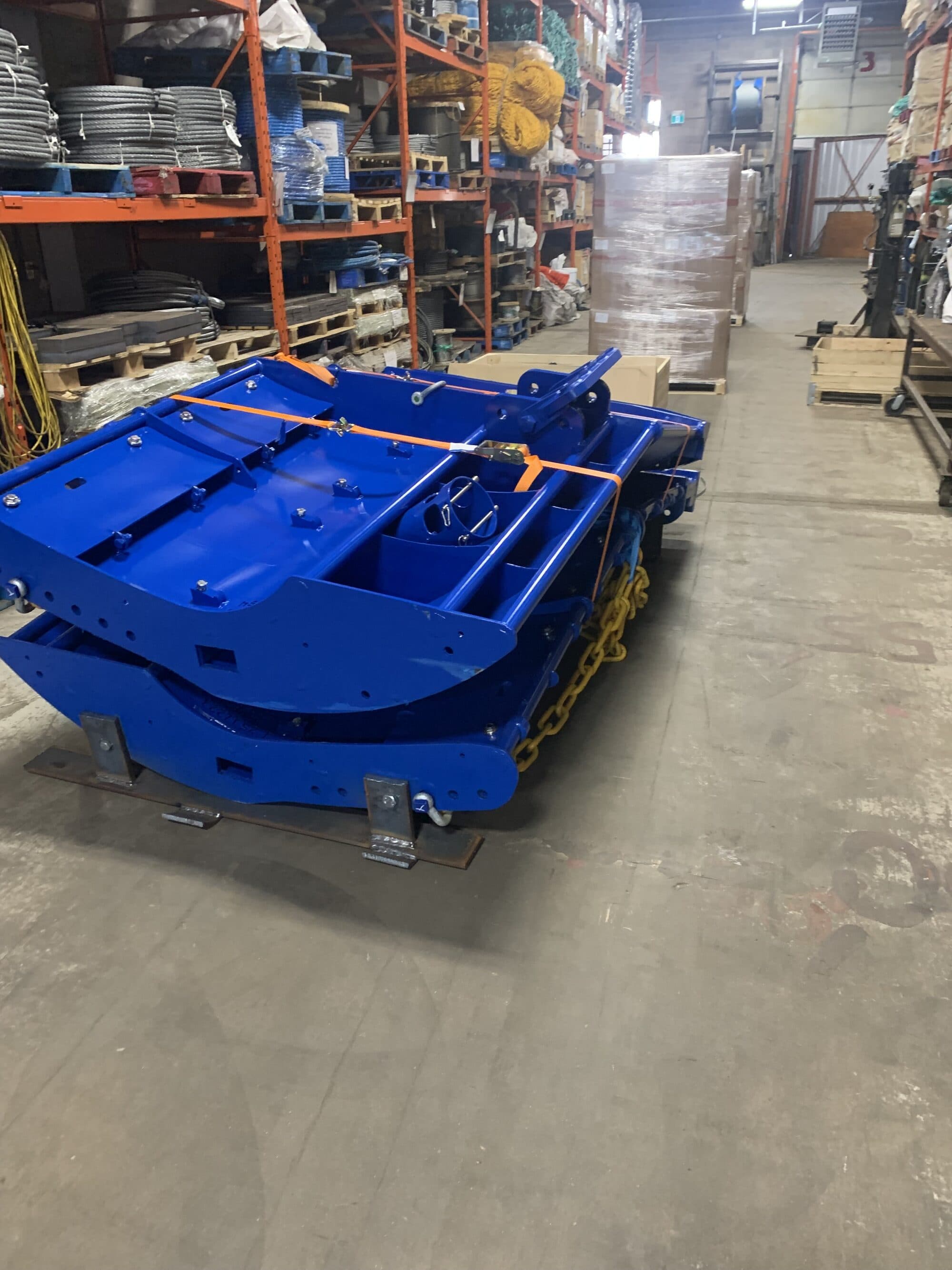Two blue Thyborøn trawl doors on the warehouse floor under an orange ratchet strap, racking of wire reels and netting behind them
