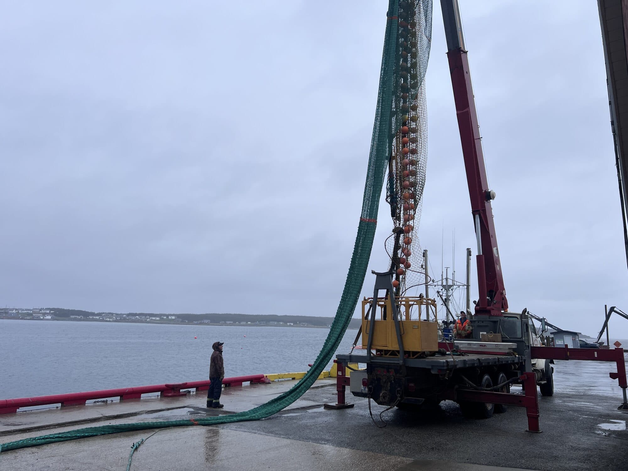 A green trawl lifted by a truck-mounted knuckle-boom crane on the wharf at Port au Choix, a crew member watching from the apron with vessels alongside