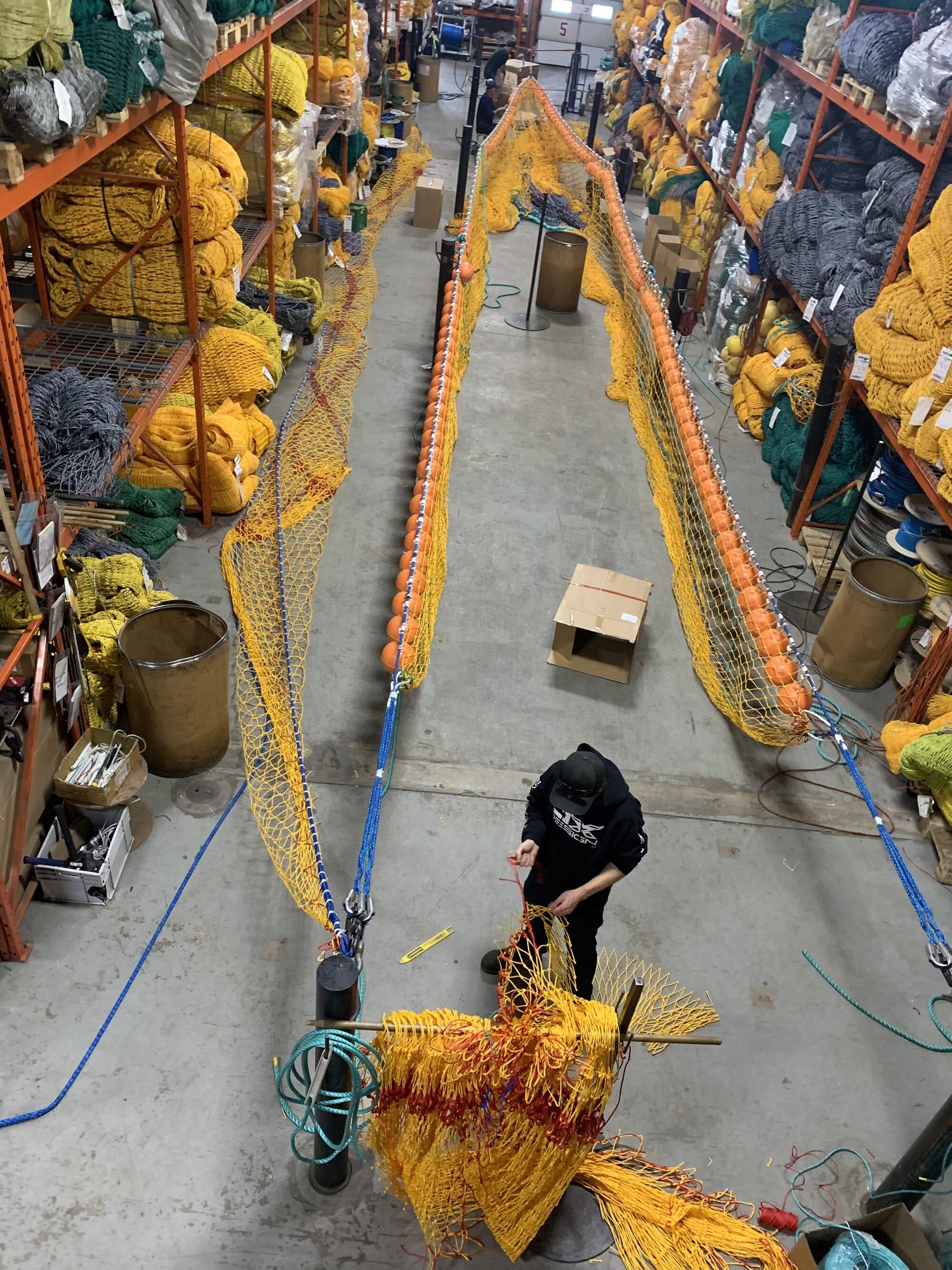 A trawl laid out the length of the loft floor with orange floats strung along its headline, a worker crouched over the yellow and red codend, netting and twine racked to both sides