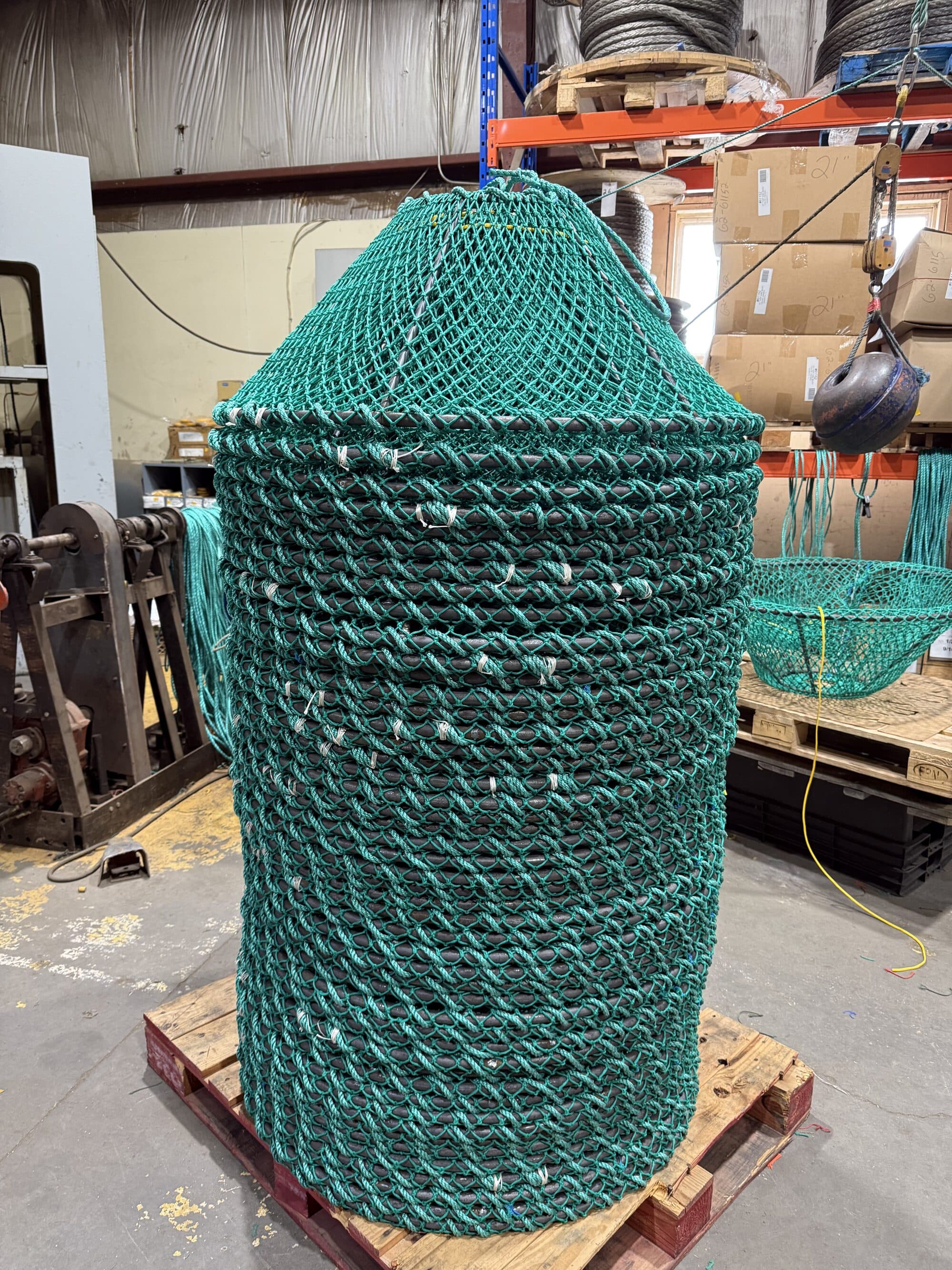 A tall stack of round pots nested on a wooden pallet in the shop, green mesh netted over each ring and seized with white twine, wire rope reels and netting racked behind
