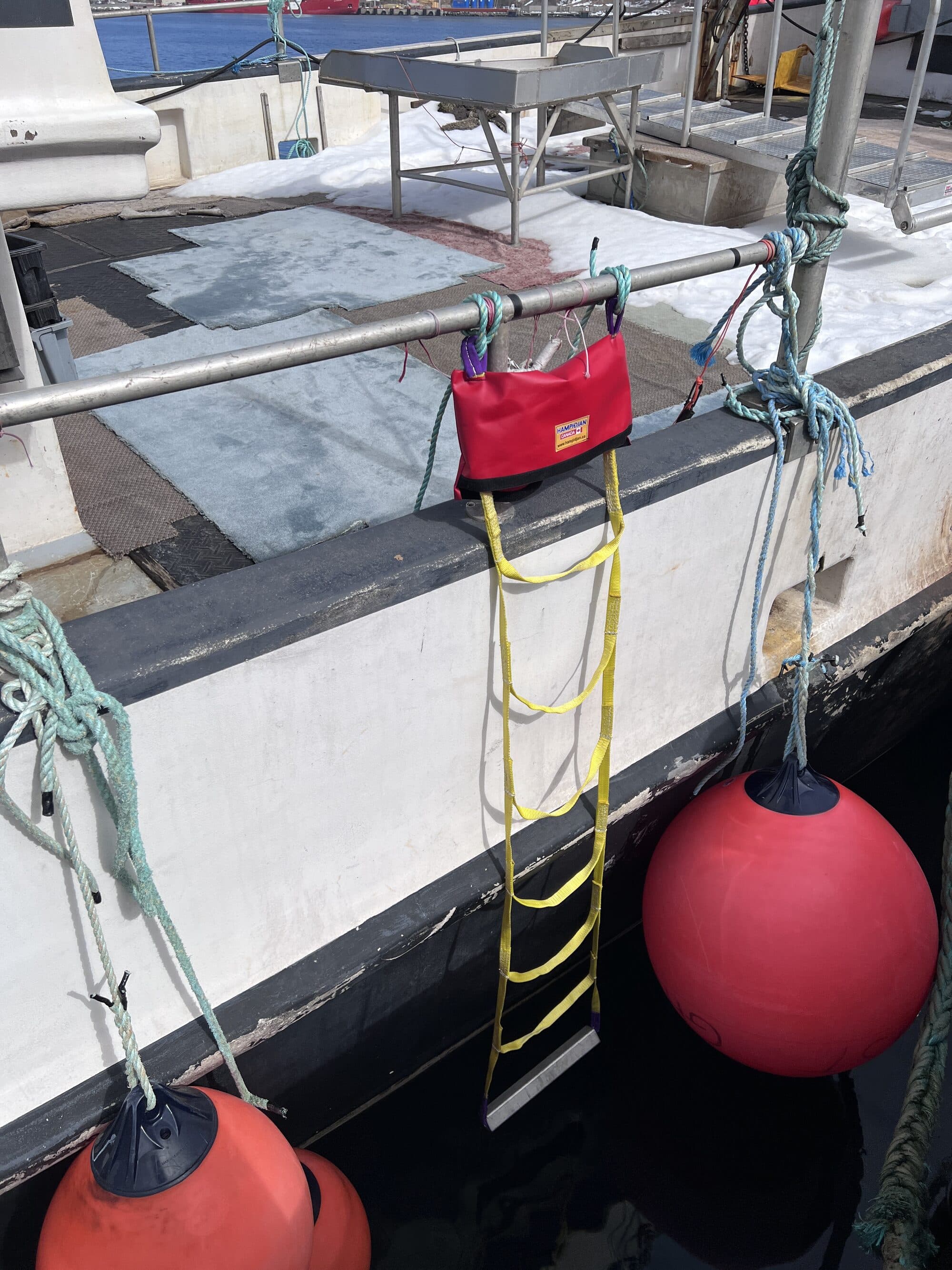 An embarkation ladder in yellow webbing with timber steps rigged over the side of a vessel and made off to the rail, fenders in the water alongside