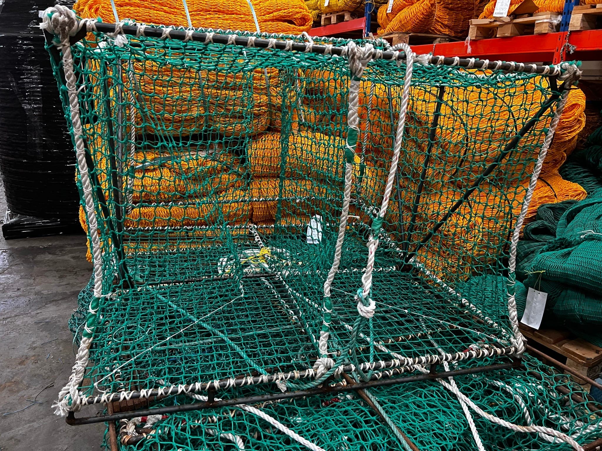 A steel frame netted in green mesh and laced with rope on the shop floor, standing among the netting stock it was built from