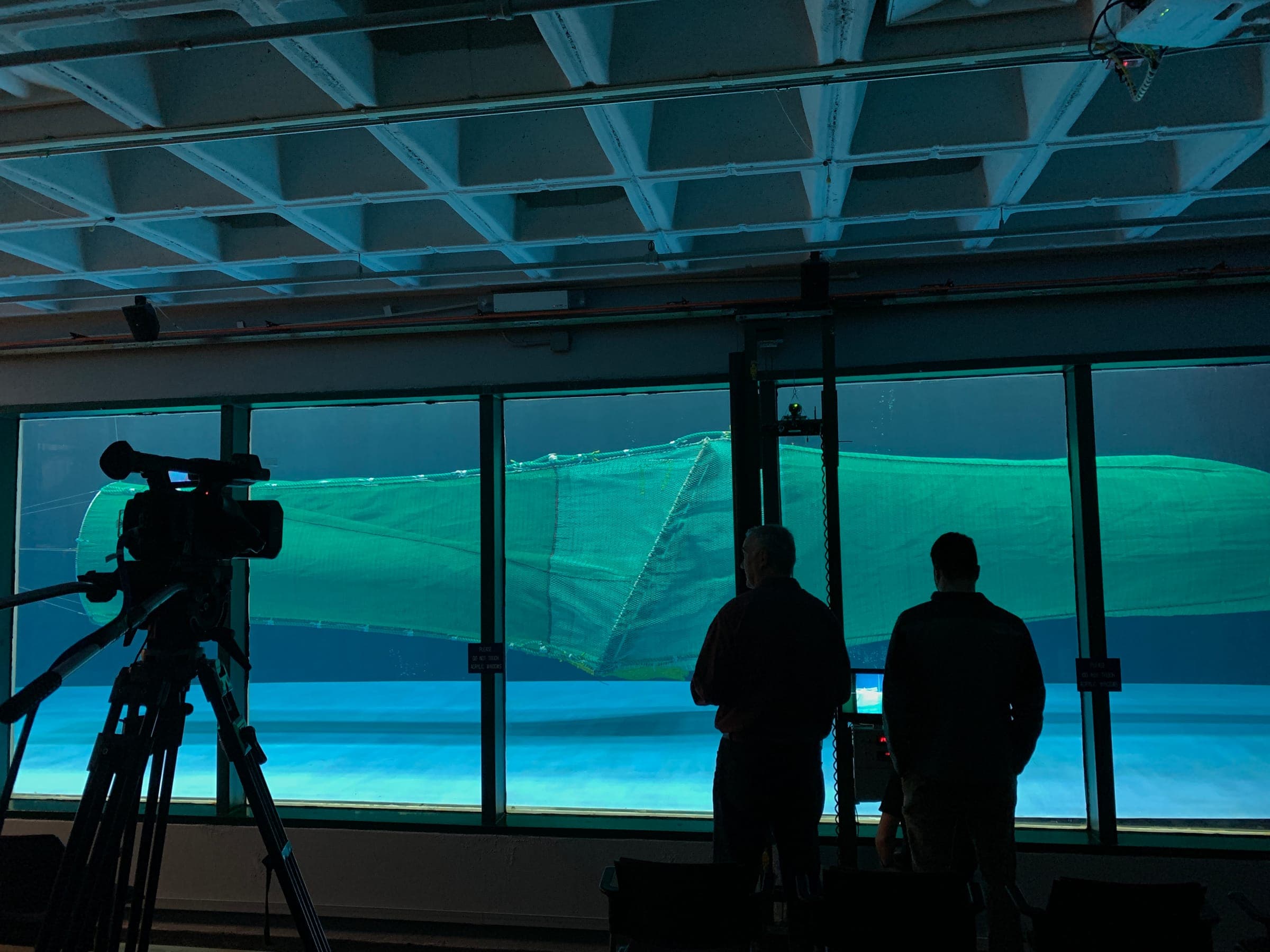 A scaled trawl model under tow in the flume tank, watched through the observation windows with a camera recording the run