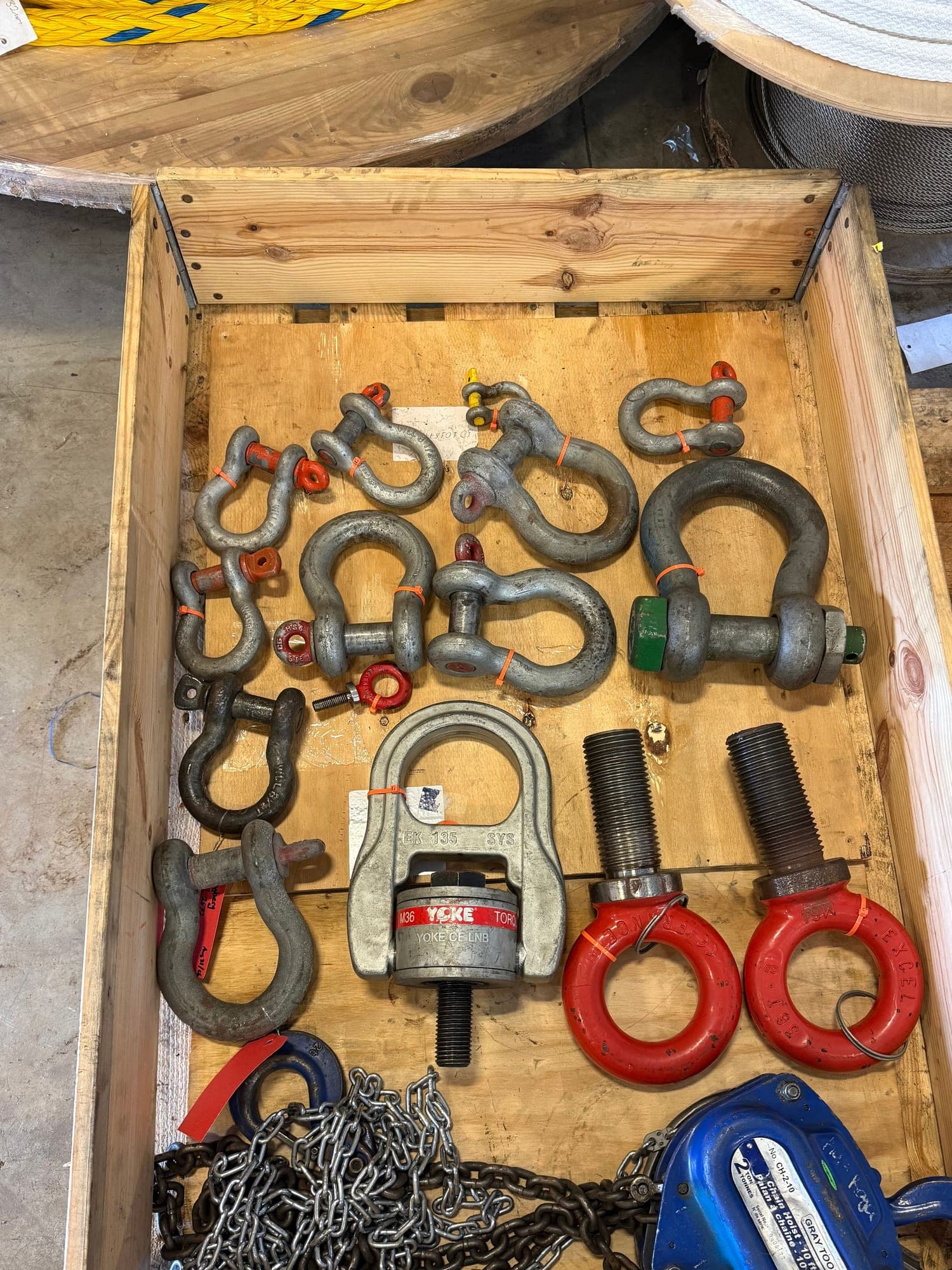 A crate of rigging hardware laid out for inspection — bow shackles in several sizes, a swivel hoist ring, eye bolts, chain and a chain hoist