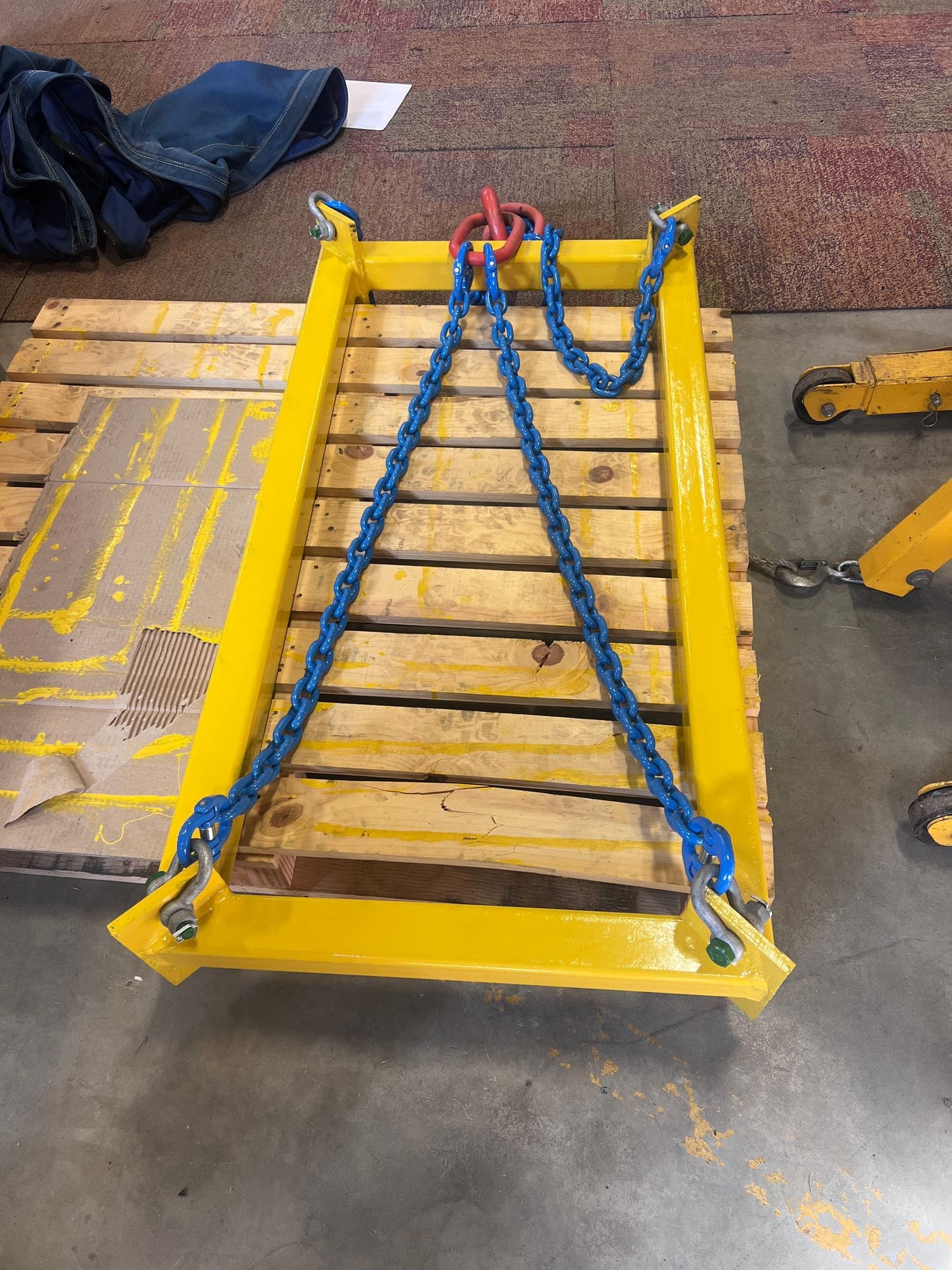 A refurbished lifting frame in fresh yellow paint with new blue chain legs, shackles at each corner and a master link at the head