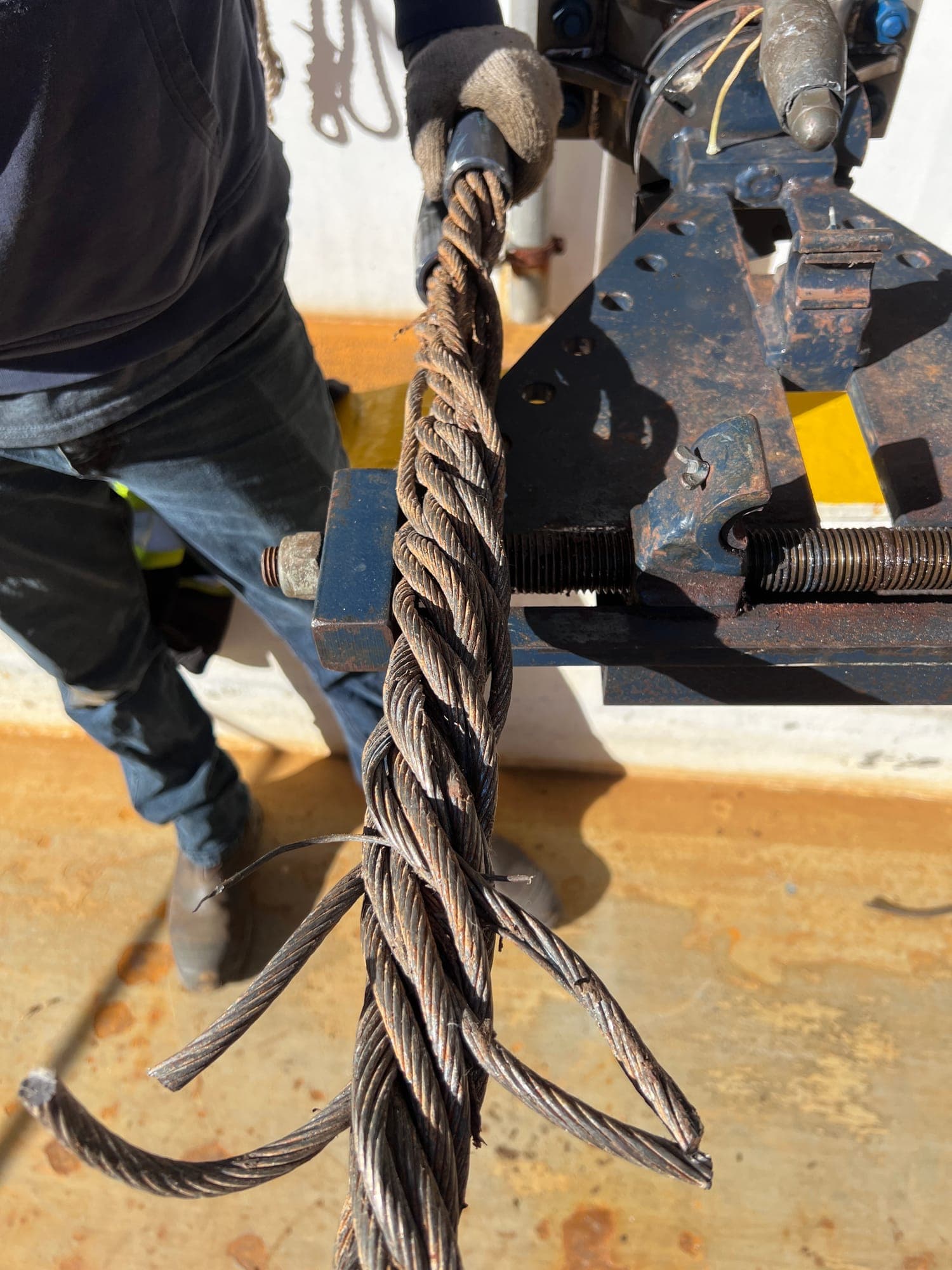 A wire rope clamped in a bench vice with its strands opened out, being spliced by hand