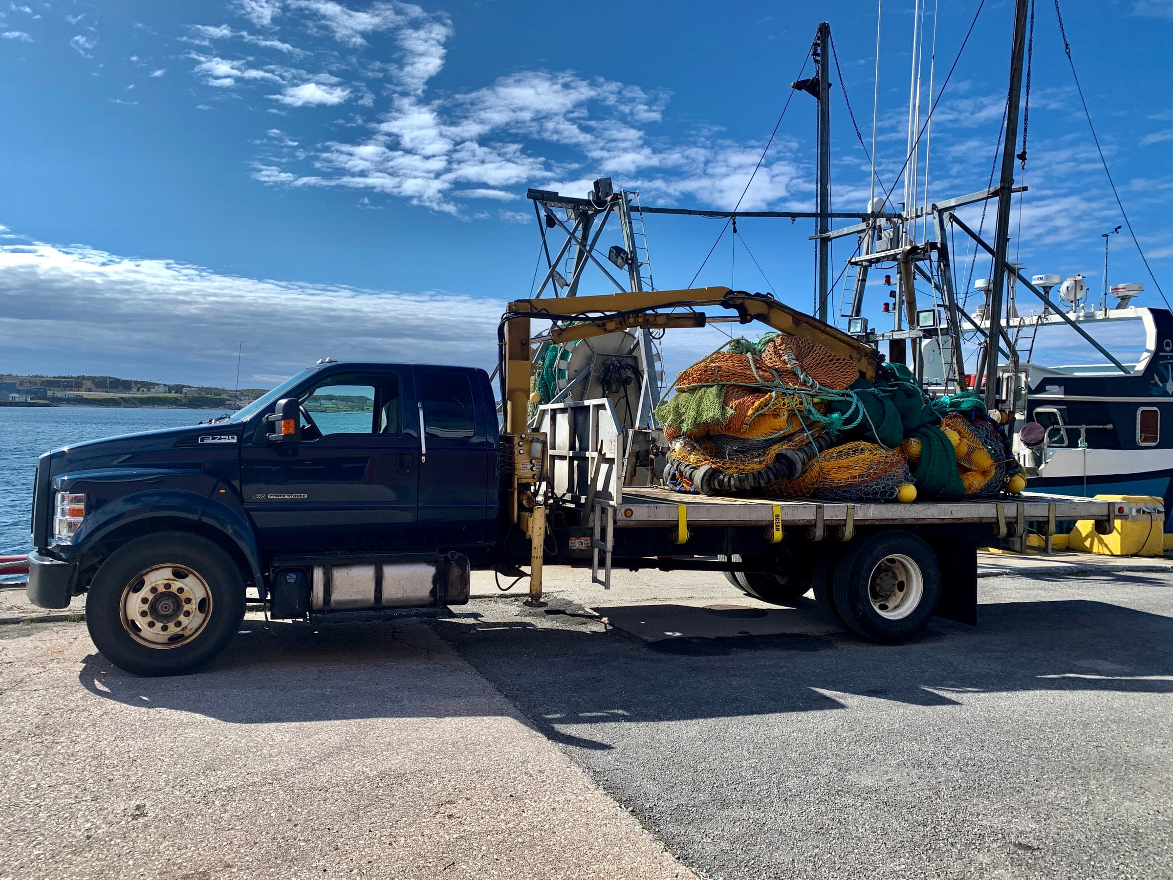 A flatbed truck with a boom crane on the wharf, loaded with a trawl in orange and green netting, a fishing vessel alongside