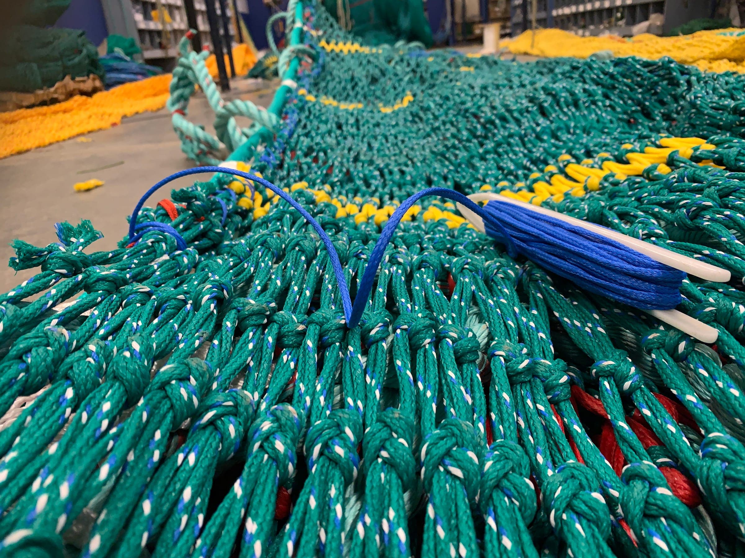 A netting needle loaded with blue twine set down in the mesh of a trawl panel part-way through a repair