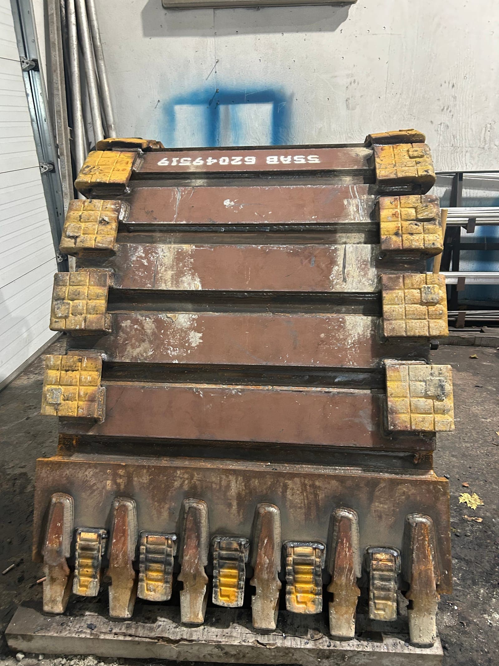 A rebuilt excavator bucket standing in the shop, new wear bars welded across its back, shrouds replaced down both sides and the teeth and adapters set along its edge