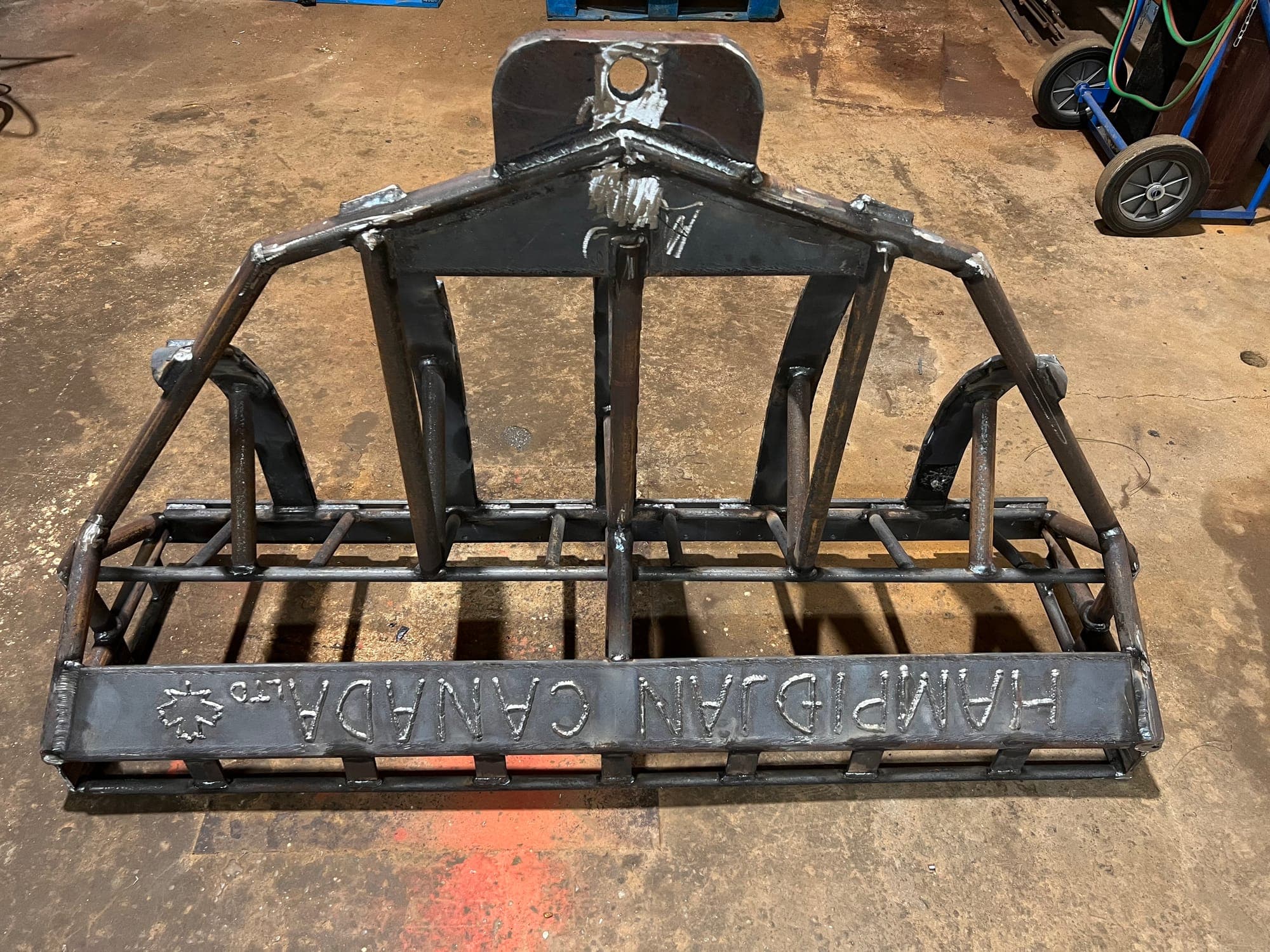 A five-bar dredge frame in bare welded steel on the shop floor, the company name welded in proud letters along its cutting bar