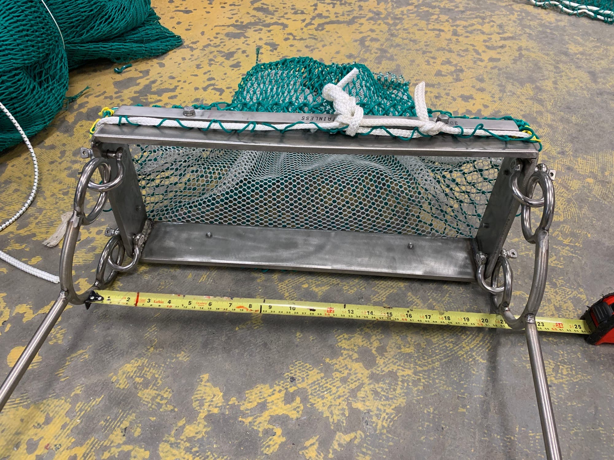 A stainless dredge frame and mesh bag on the shop floor with a tape measure laid across the build