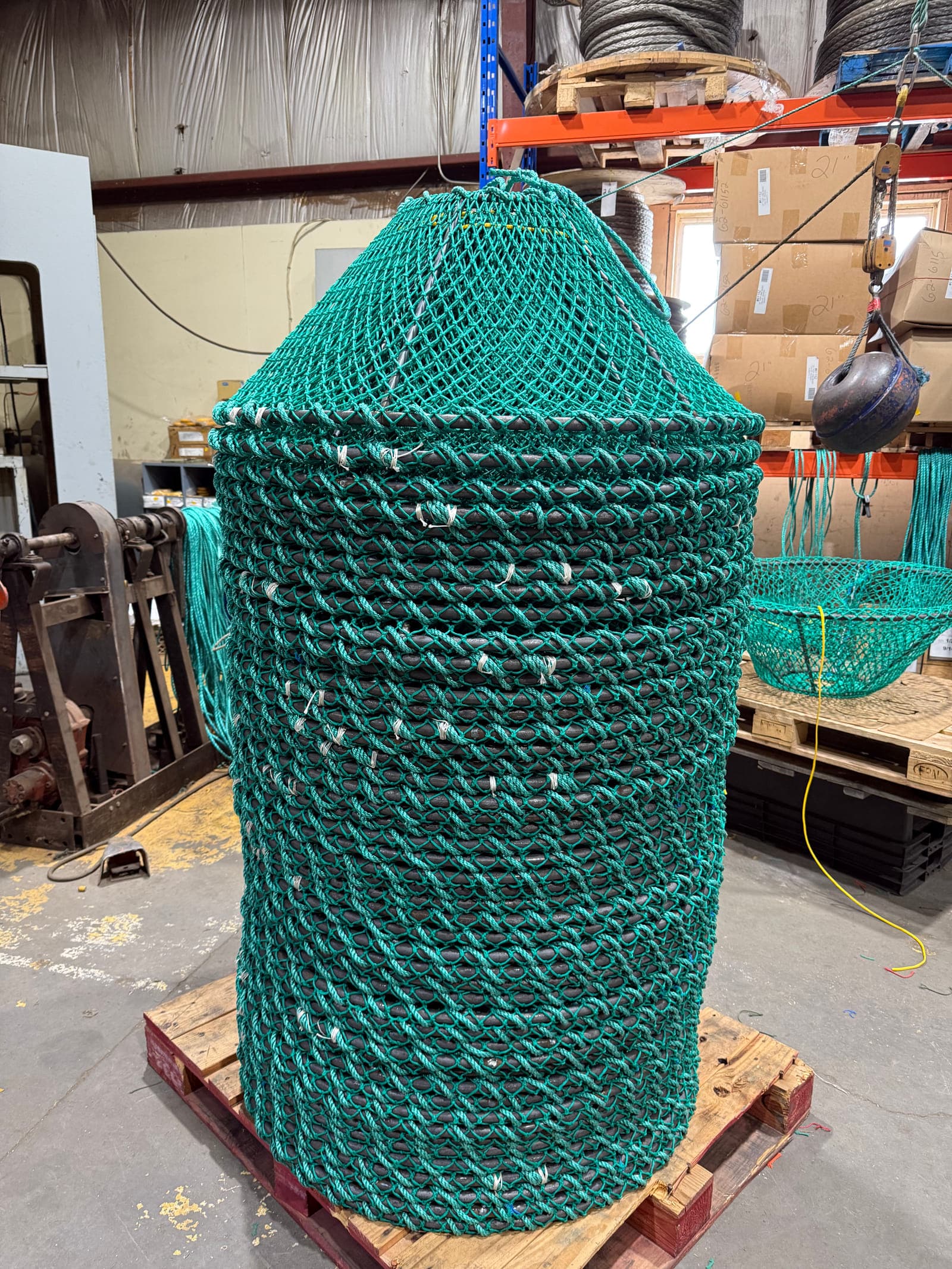 A stack of netted crab pots on a wooden pallet in the shop, green twine laced over the steel frames, wire reels racked behind